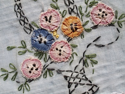 Intricately embroidered flowers in various colors are sewn onto a light fabric. The flowers include shades of pink, blue, and orange, each with detailed stitching and black accents resembling seeds or pollen. Green leaves and stems are also embroidered, adding a natural element to the design.