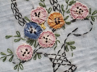 Intricately embroidered flowers in various colors are sewn onto a light fabric. The flowers include shades of pink, blue, and orange, each with detailed stitching and black accents resembling seeds or pollen. Green leaves and stems are also embroidered, adding a natural element to the design.