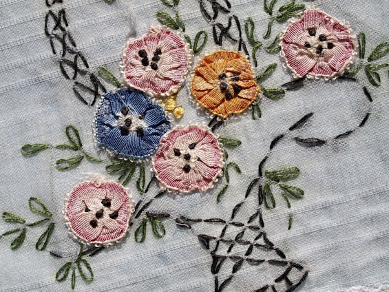 Intricately embroidered flowers in various colors are sewn onto a light fabric. The flowers include shades of pink, blue, and orange, each with detailed stitching and black accents resembling seeds or pollen. Green leaves and stems are also embroidered, adding a natural element to the design.