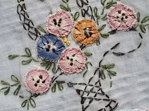 Intricately embroidered flowers in various colors are sewn onto a light fabric. The flowers include shades of pink, blue, and orange, each with detailed stitching and black accents resembling seeds or pollen. Green leaves and stems are also embroidered, adding a natural element to the design.