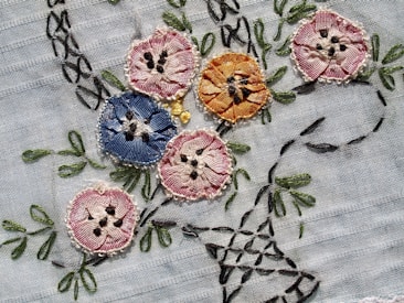 Intricately embroidered flowers in various colors are sewn onto a light fabric. The flowers include shades of pink, blue, and orange, each with detailed stitching and black accents resembling seeds or pollen. Green leaves and stems are also embroidered, adding a natural element to the design.