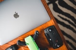 A silver laptop with an Apple logo, a green and black drone, and a black remote control are placed on a wooden table with a zebra-striped patterned rug partially visible in the background.