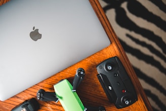 A silver laptop with an Apple logo, a green and black drone, and a black remote control are placed on a wooden table with a zebra-striped patterned rug partially visible in the background.