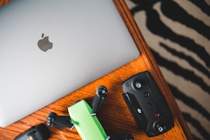 A silver laptop with an Apple logo, a green and black drone, and a black remote control are placed on a wooden table with a zebra-striped patterned rug partially visible in the background.