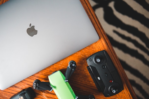 A silver laptop with an Apple logo, a green and black drone, and a black remote control are placed on a wooden table with a zebra-striped patterned rug partially visible in the background.