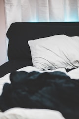 A bed with white sheets and a black blanket, featuring a white pillow resting against a black headboard. The background includes white curtains.