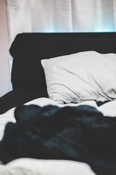 A bed with white sheets and a black blanket, featuring a white pillow resting against a black headboard. The background includes white curtains.