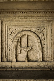 A detailed stone carving featuring an ornate arched design. The carving includes intricate patterns and motifs around the central arch, displaying a combination of floral and geometric elements. The texture of the stone appears aged and weathered, highlighting the craftsmanship involved.