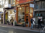 A streetscape featuring a row of shops with varied architectural styles and large windows. The storefronts include a coffee shop and an Indian kitchen restaurant. Bicycles are parked along the sidewalk, and two men are walking by carrying shopping bags.