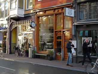 A streetscape featuring a row of shops with varied architectural styles and large windows. The storefronts include a coffee shop and an Indian kitchen restaurant. Bicycles are parked along the sidewalk, and two men are walking by carrying shopping bags.
