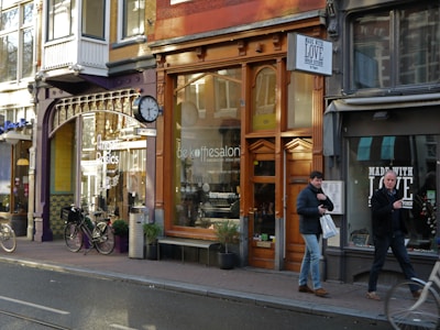 A streetscape featuring a row of shops with varied architectural styles and large windows. The storefronts include a coffee shop and an Indian kitchen restaurant. Bicycles are parked along the sidewalk, and two men are walking by carrying shopping bags.