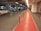 An indoor parking garage with several parked cars. The floor is marked with a red path separated by orange bollards, leading towards the exit. The ceiling is equipped with lighting and pipes. Two people are walking along the red path towards the exit. The walls have informational signs, and the area is well-lit.