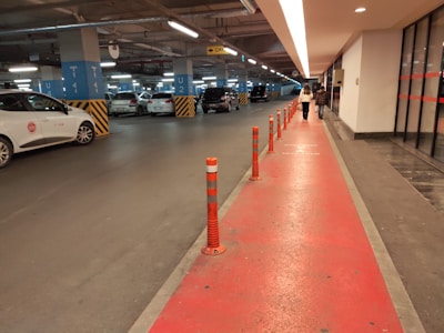 An indoor parking garage with several parked cars. The floor is marked with a red path separated by orange bollards, leading towards the exit. The ceiling is equipped with lighting and pipes. Two people are walking along the red path towards the exit. The walls have informational signs, and the area is well-lit.