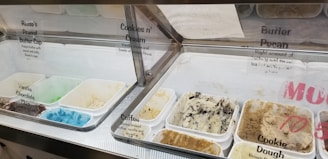 An assortment of ice cream flavors is displayed in a freezer case, with clear containers holding various ice cream types such as Reese's Peanut Butter Cup, Cookies n' Cream, Vanilla, Chocolate, Coffee, and Cookie Dough. The ice creams range in color from bright blue to muted browns and creams. Each flavor has a label with the name and a brief description.