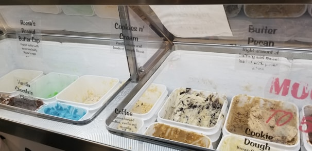 An assortment of ice cream flavors is displayed in a freezer case, with clear containers holding various ice cream types such as Reese's Peanut Butter Cup, Cookies n' Cream, Vanilla, Chocolate, Coffee, and Cookie Dough. The ice creams range in color from bright blue to muted browns and creams. Each flavor has a label with the name and a brief description.