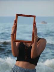 A person stands on a beach holding a wooden rectangular frame in front of their face, creating the illusion of invisibility. The ocean waves in the background add to the surreal composition. The person is wearing a black top and blue patterned pants, with a cross necklace and a watch.
