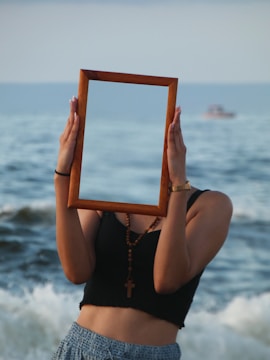 A person stands on a beach holding a wooden rectangular frame in front of their face, creating the illusion of invisibility. The ocean waves in the background add to the surreal composition. The person is wearing a black top and blue patterned pants, with a cross necklace and a watch.