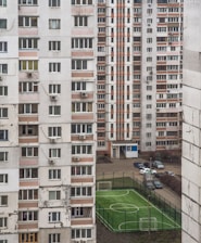 A perspective view of residential apartment buildings with numerous windows and balconies, characterized by a mix of white and faded orange panels. Between the buildings, a small fenced sports field with two goalposts is visible, and several cars are parked nearby.
