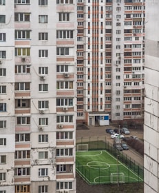 A perspective view of residential apartment buildings with numerous windows and balconies, characterized by a mix of white and faded orange panels. Between the buildings, a small fenced sports field with two goalposts is visible, and several cars are parked nearby.
