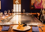A Japanese teppanyaki grill setup is illustrated, featuring a central metallic cooking surface surrounded by wooden countertops. The table is set with wine glasses, blue napkins, and cutlery. In the foreground, there is a plate of tempura with dipping sauce. The background showcases a panel with illuminated Japanese Kanji characters and an artistic wall decoration.