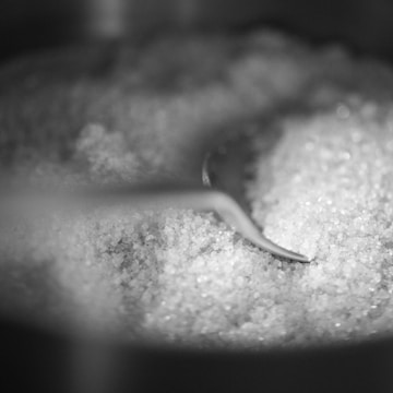 A pile of granulated sugar with a metal spoon partially buried in it. The texture of the sugar crystals is highlighted, creating a granular and shiny appearance.