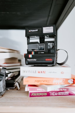 A vintage Polaroid camera rests on a stack of books. The books include 'Invisible Man' by Ralph Ellison, an orange book, and a book by Jon Ronson titled 'Them: Adventures with Extremists'. The scene is set on a wooden surface with a dark, out-of-focus background.