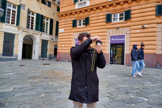 A person in a black coat is taking a photograph in an urban area with a stone-paved street. The background features an orange building with green shuttered windows and a store entrance labeled 'Marionnaud'. Two other individuals wearing winter jackets and jeans are walking by.