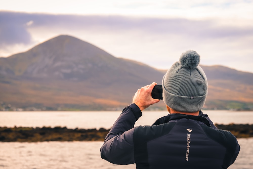 Hombre con chaqueta negra y gorra de punto gris tomando foto de la montaña durante el día