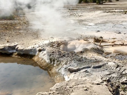 A geothermal area with steam rising from the ground, surrounded by rocky terrain and pools of water. The surface is uneven and crusted with mineral deposits, creating a barren, otherworldly landscape.