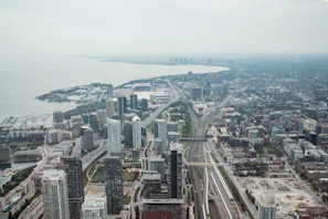 Aerial view of a modern cityscape featuring numerous high-rise buildings and dense urban infrastructure. The wide boulevard and railway tracks extend into the distance, leading towards a large body of water. The city is surrounded by greenery and residential areas stretching towards the horizon.