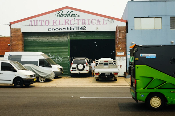 A garage with a sign reading 'Auto Electrical' above the entrance. Several vehicles, including a white van and a flatbed truck, are parked in front of the building. A person is seen working near the vehicles. A green street sweeper or council vehicle is on the street in the foreground.