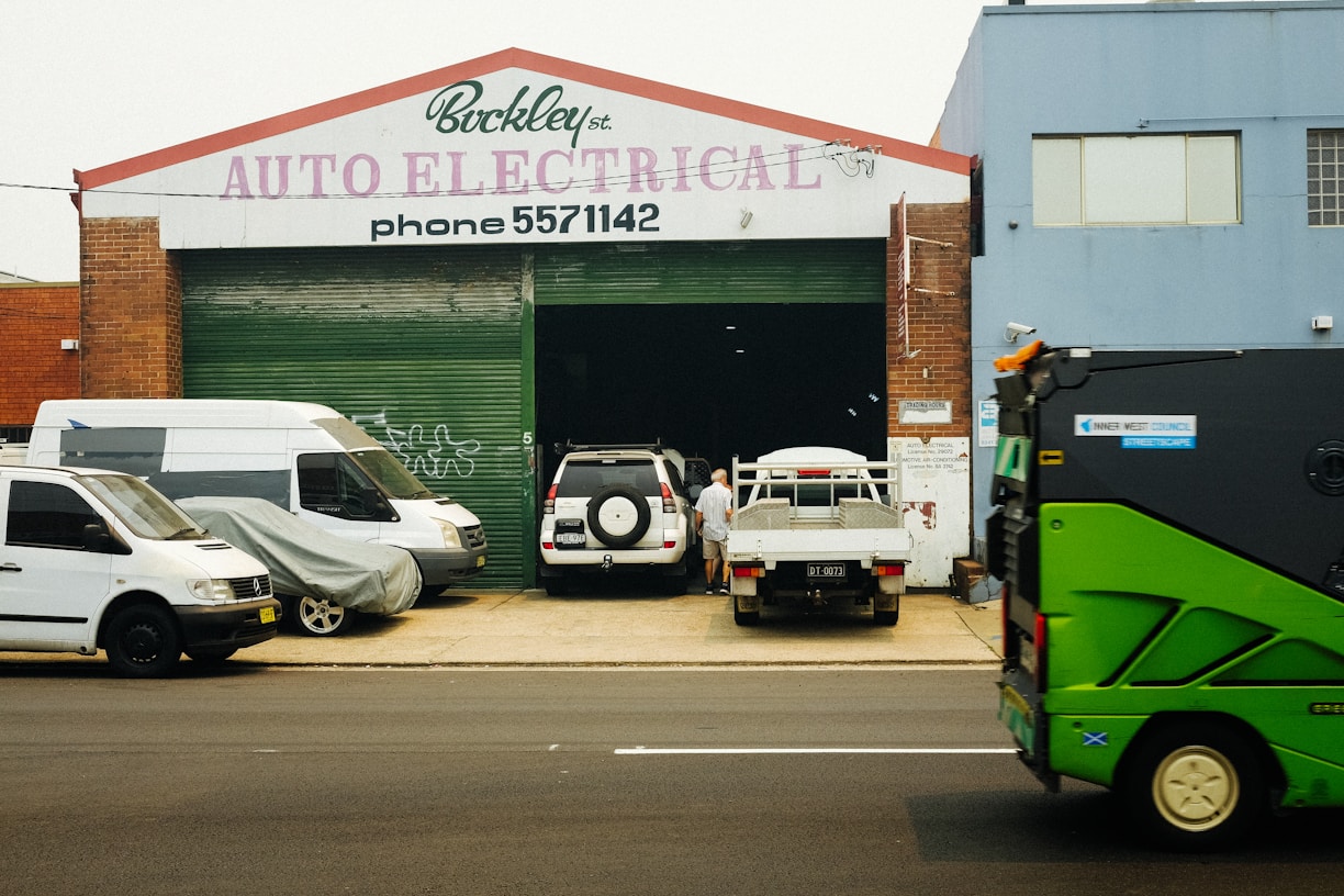 A garage with a sign reading 'Auto Electrical' above the entrance. Several vehicles, including a white van and a flatbed truck, are parked in front of the building. A person is seen working near the vehicles. A green street sweeper or council vehicle is on the street in the foreground.