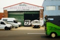 A garage with a sign reading 'Auto Electrical' above the entrance. Several vehicles, including a white van and a flatbed truck, are parked in front of the building. A person is seen working near the vehicles. A green street sweeper or council vehicle is on the street in the foreground.