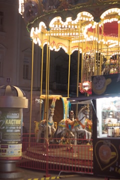 A brightly lit carousel with ornate white and gold decorations and several colorful horses is situated in an outdoor setting. The carousel's lights create a warm glow in the surrounding area. Nearby, there is a vending stall with various items for sale and a tall advertisement pillar.