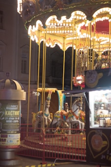 A brightly lit carousel with ornate white and gold decorations and several colorful horses is situated in an outdoor setting. The carousel's lights create a warm glow in the surrounding area. Nearby, there is a vending stall with various items for sale and a tall advertisement pillar.