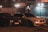 Two cars are parked next to each other at night under streetlights. One is an older model with a darker color, while the other is a modern white sedan. The background features a building with an illuminated sign and metal fencing, contributing to an urban nighttime atmosphere.