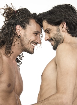 Two men are standing closely, smiling warmly at each other with their foreheads touching. Both appear to be topless, emphasizing a sense of closeness and intimacy.