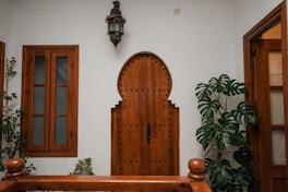A wooden door with an arched design, featuring decorative studs, is centered between two wooden-framed windows with frosted glass panes. A large leafy plant stands to the right of the door, and an ornate lantern hangs above. The walls are painted white, creating a clean, minimalist look.