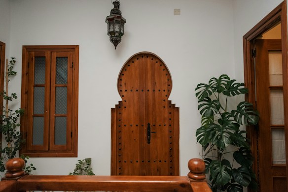 A wooden door with an arched design, featuring decorative studs, is centered between two wooden-framed windows with frosted glass panes. A large leafy plant stands to the right of the door, and an ornate lantern hangs above. The walls are painted white, creating a clean, minimalist look.