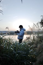 A person, holding a camera, stands amidst tall grasses with a serene backdrop of a misty lake at sunrise. The sunlight creates a soft glow around the person, highlighting the peaceful and reflective atmosphere.