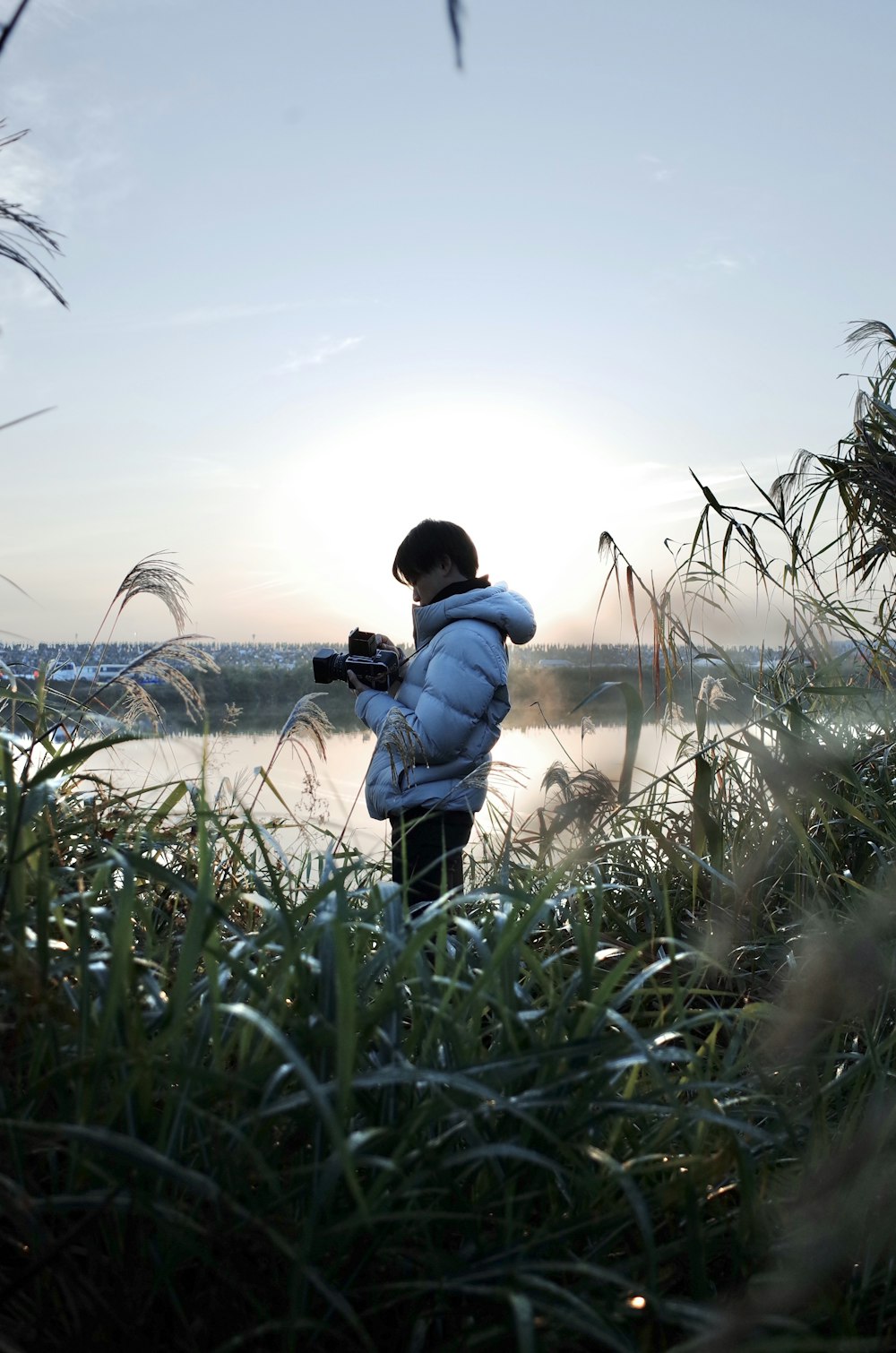 青いジャケットと黒いズボンを着た男が、緑の芝生の野原に立つ黒いデジタル一眼レフカメラを手にしている