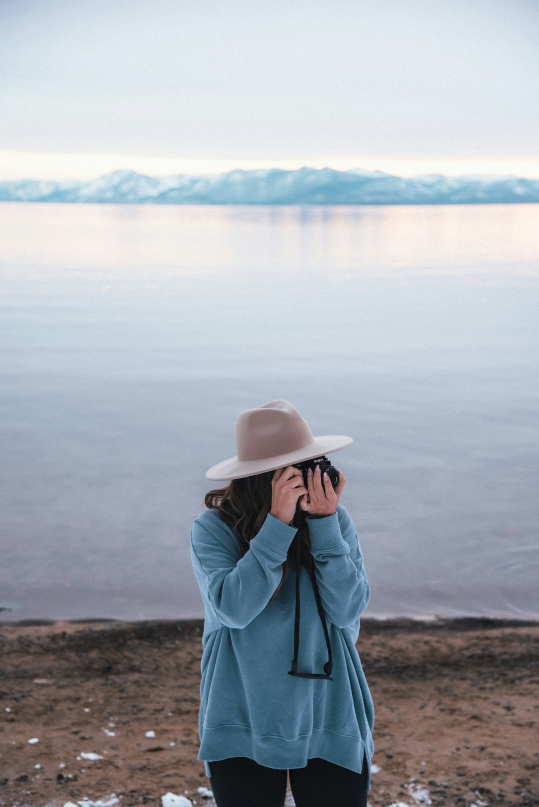 woman in blue hoodie wearing white fedora hat standing on brown sand near body of water