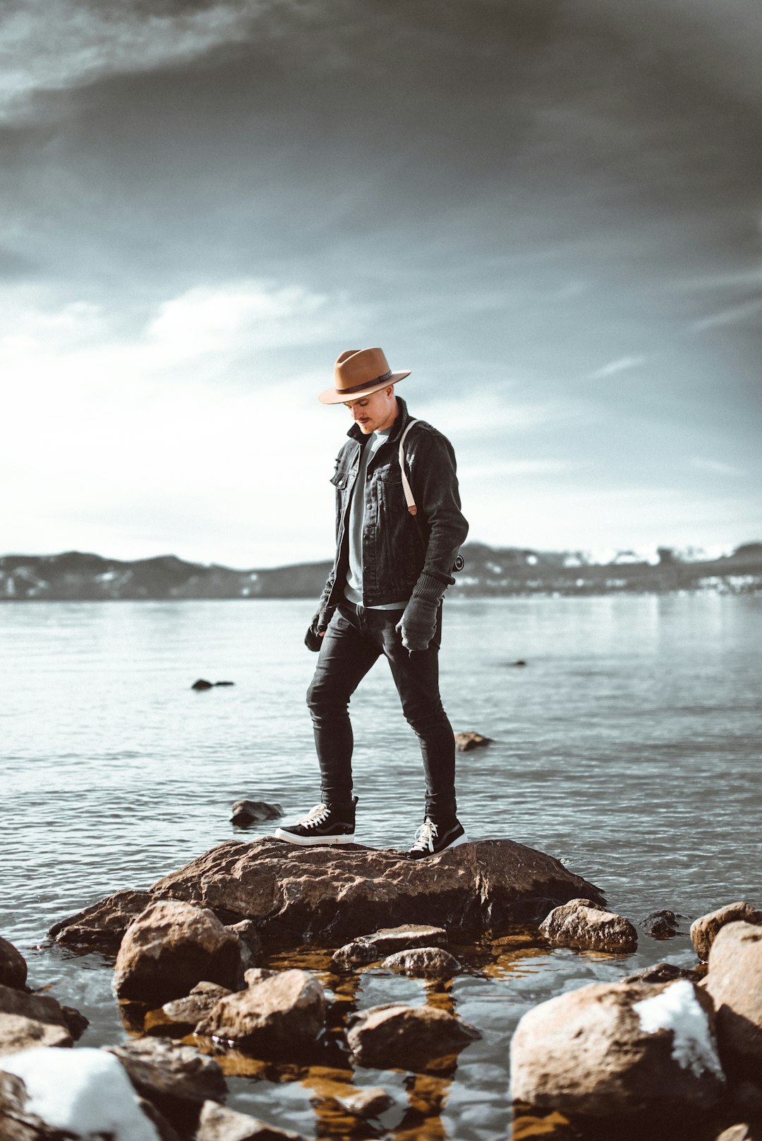 man in black jacket and brown hat standing on brown rock near body of water during