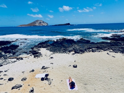 A person is sunbathing on a towel spread on a sandy beach, with a clear blue sea and rocky shoreline in the background. An island is visible on the horizon under a bright sky scattered with a few clouds.