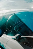 A close-up view through the windshield of a car, focusing on the steering wheel and dashboard area. The glass reflects light, creating a slightly distorted view of the interior.