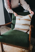 A person is arranging a decorative cushion on a green upholstered armchair. The cushion has a pattern of white and brown stripes. The chair has wooden armrests and is placed on a floor with a light brown rug.