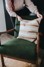 A person is arranging a decorative cushion on a green upholstered armchair. The cushion has a pattern of white and brown stripes. The chair has wooden armrests and is placed on a floor with a light brown rug.