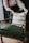 A person is arranging a decorative cushion on a green upholstered armchair. The cushion has a pattern of white and brown stripes. The chair has wooden armrests and is placed on a floor with a light brown rug.
