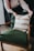 A person is arranging a decorative cushion on a green upholstered armchair. The cushion has a pattern of white and brown stripes. The chair has wooden armrests and is placed on a floor with a light brown rug.
