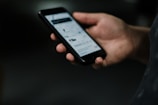A hand is holding a smartphone with the screen displaying a webpage or app interface, featuring text and images. The background is dark, highlighting the phone.